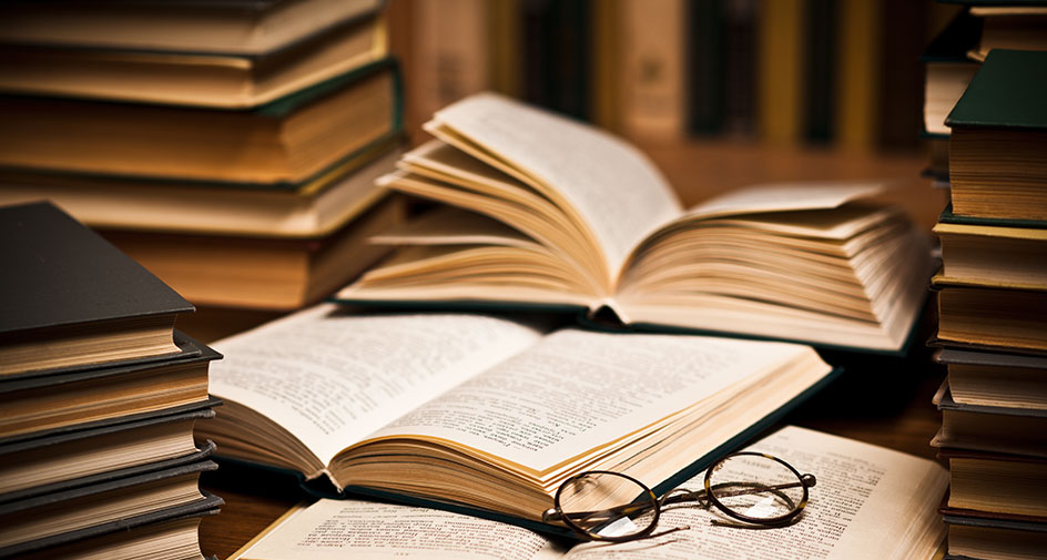 Two stacks of aged looking books with an open book with a pair of reading glasses on top laying between them.